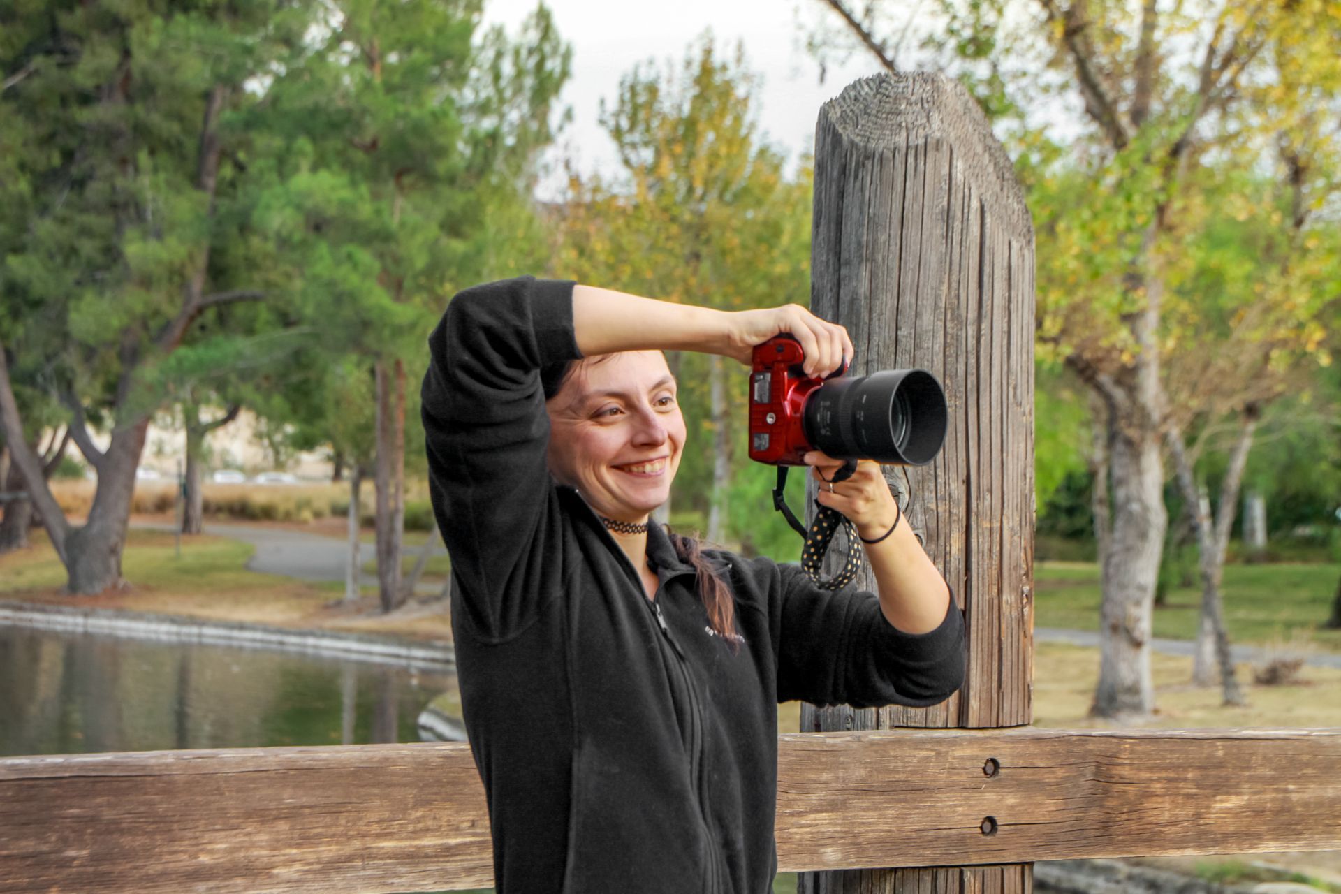 Woman taking a photo with a red camera. She smiles, standing near a wooden fence and trees by a pond.