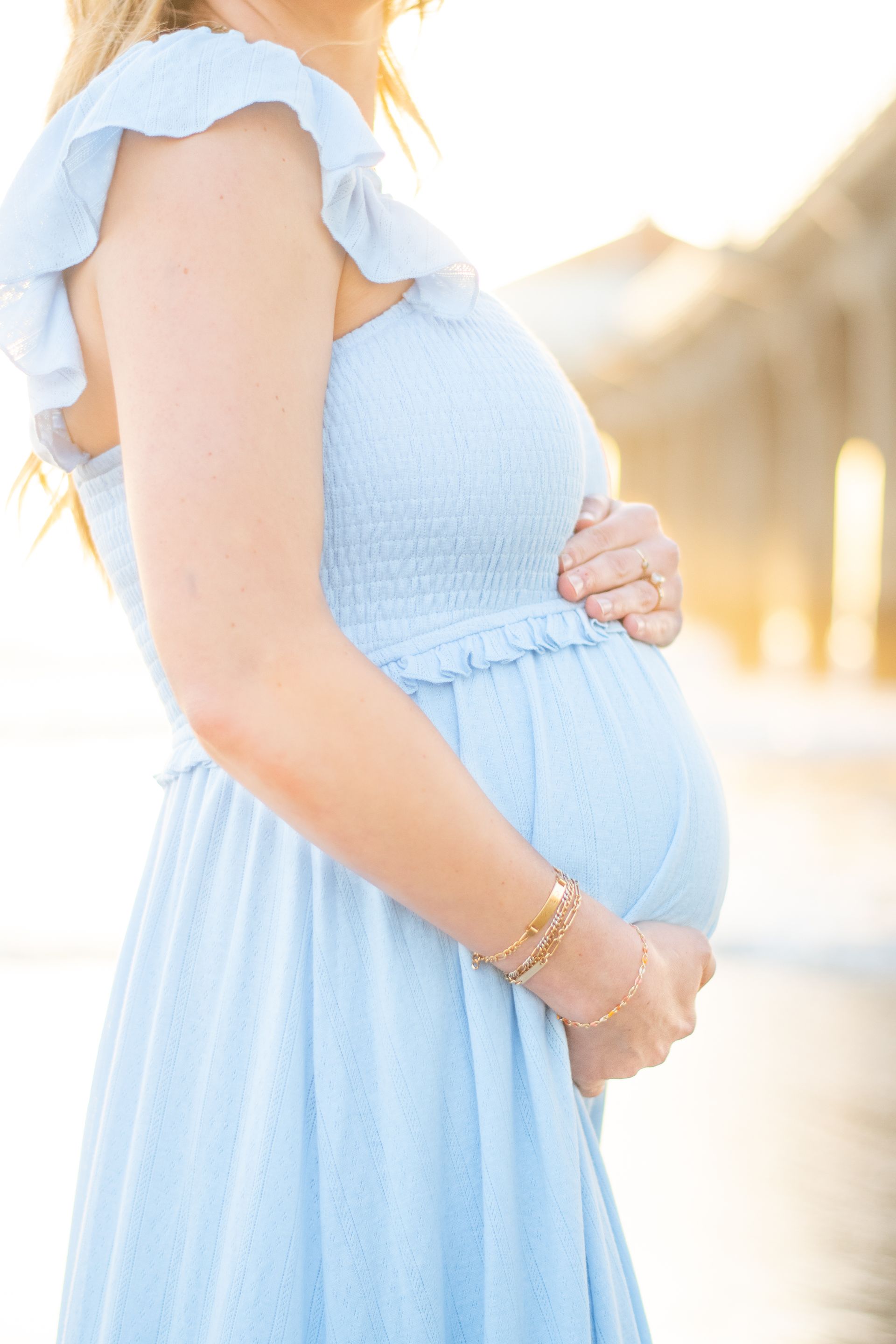 Pregnant woman in blue dress, hands on her baby bump, near a pier at sunset.