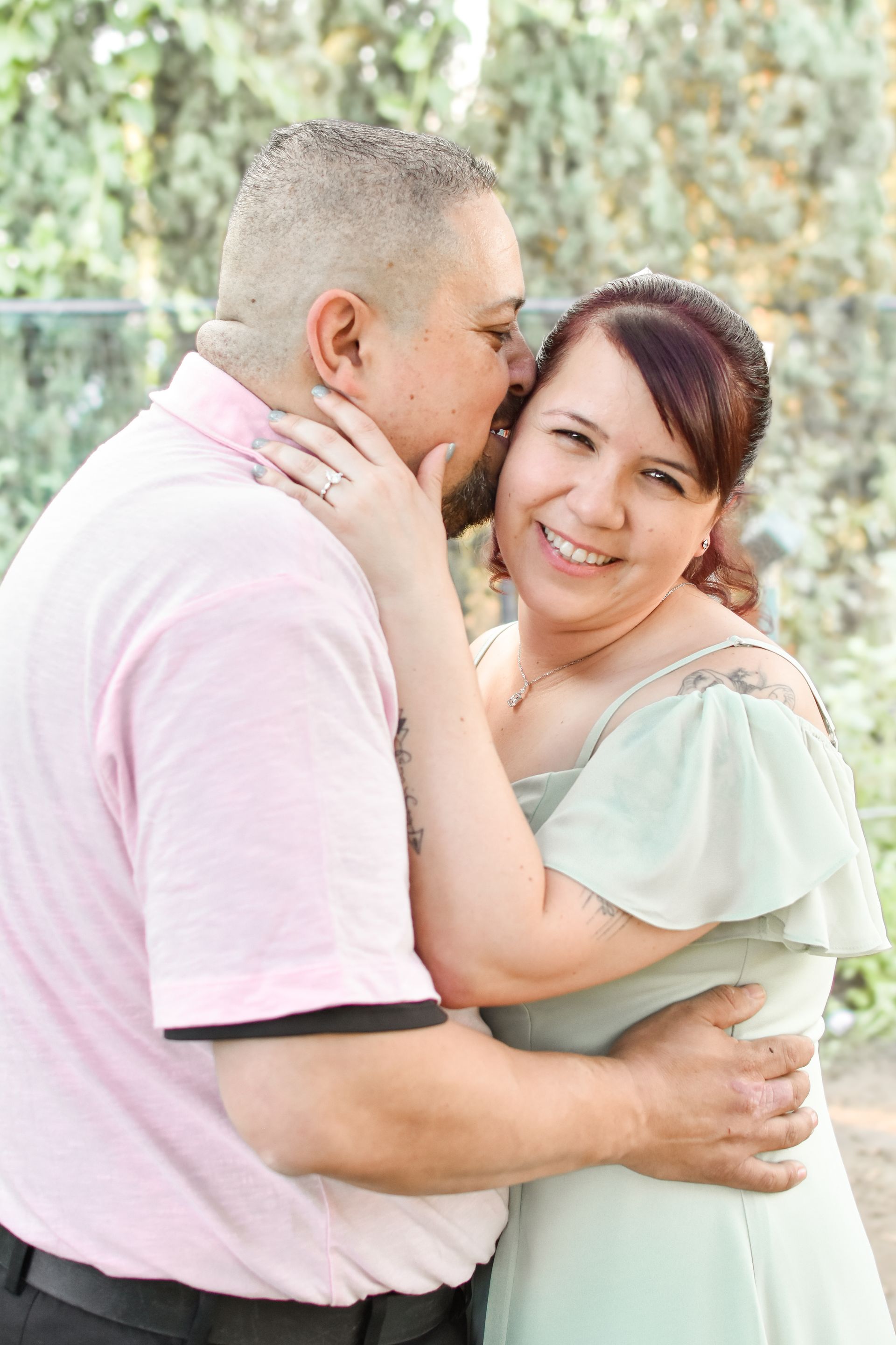 A person in a light pink polo shirt kisses a person in a light green dress on the cheek, outdoors in a park setting.