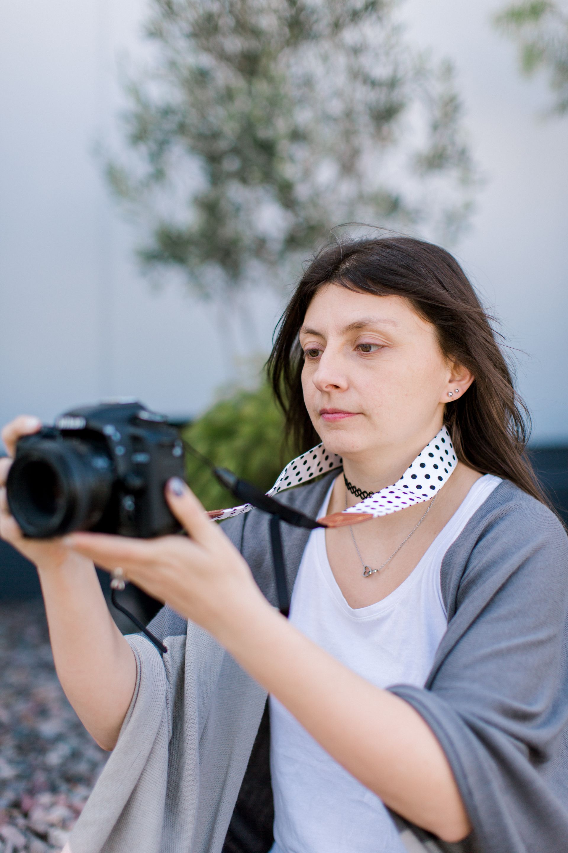 Woman holding a camera, looking at the screen. She is outdoors, wearing a white shirt and gray shawl.