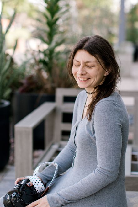 Woman in gray dress sits smiling, holding camera outdoors.