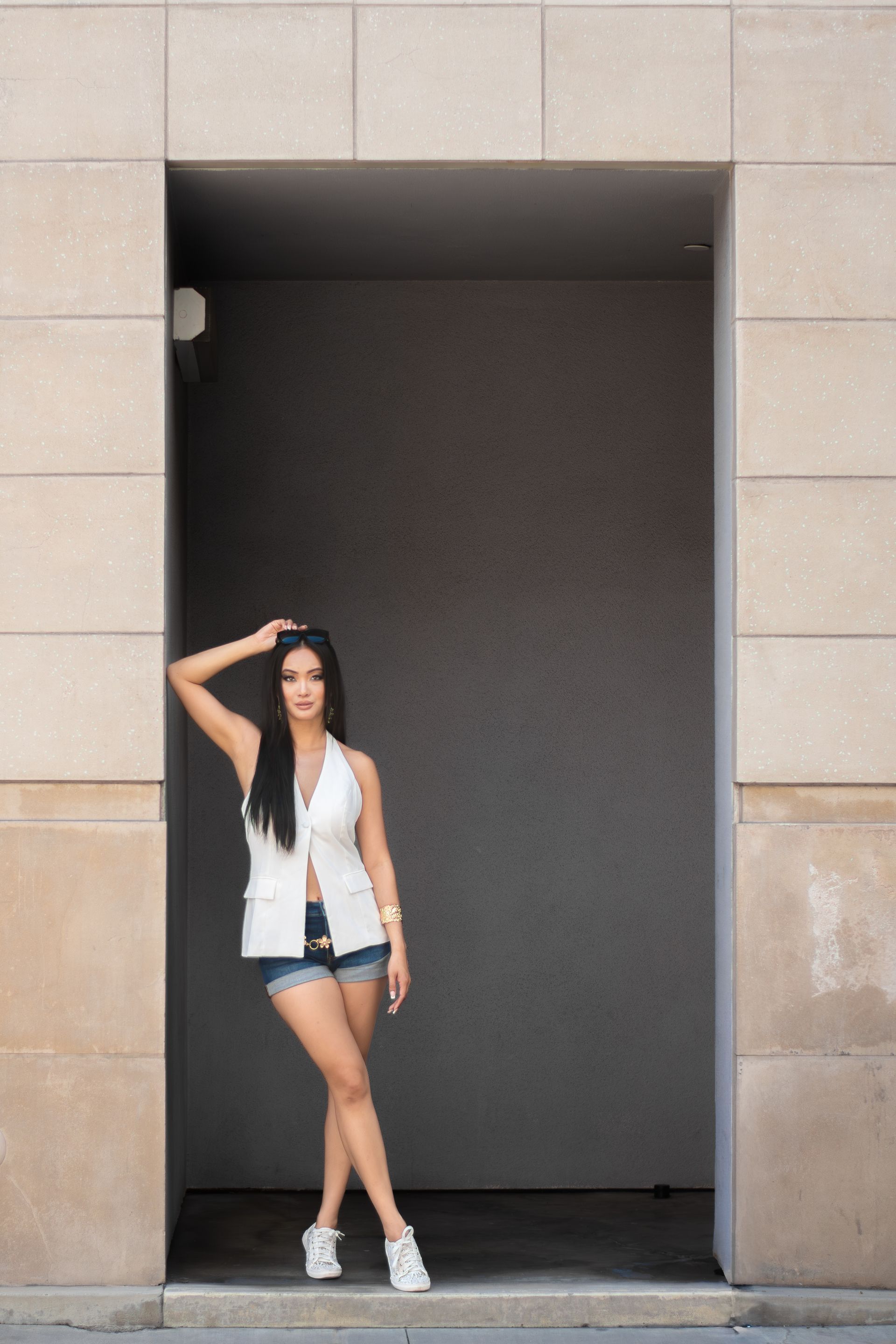 Woman standing in a doorway, wearing a white top, denim shorts, and sneakers, with one hand on her head.