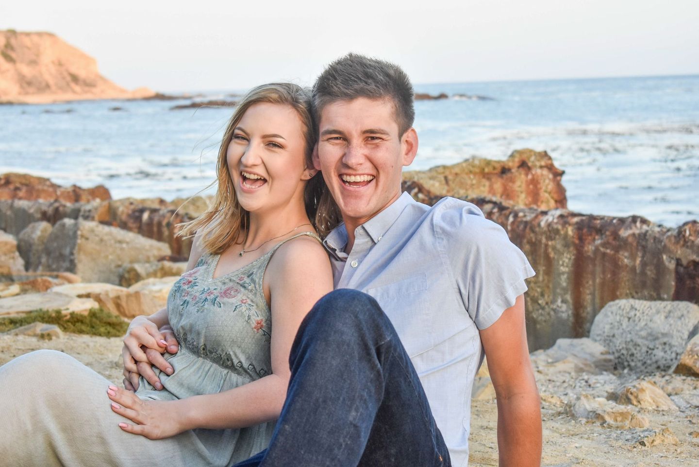 Couple laughing, seated on rocks by the ocean; woman in floral dress, man in blue shirt.