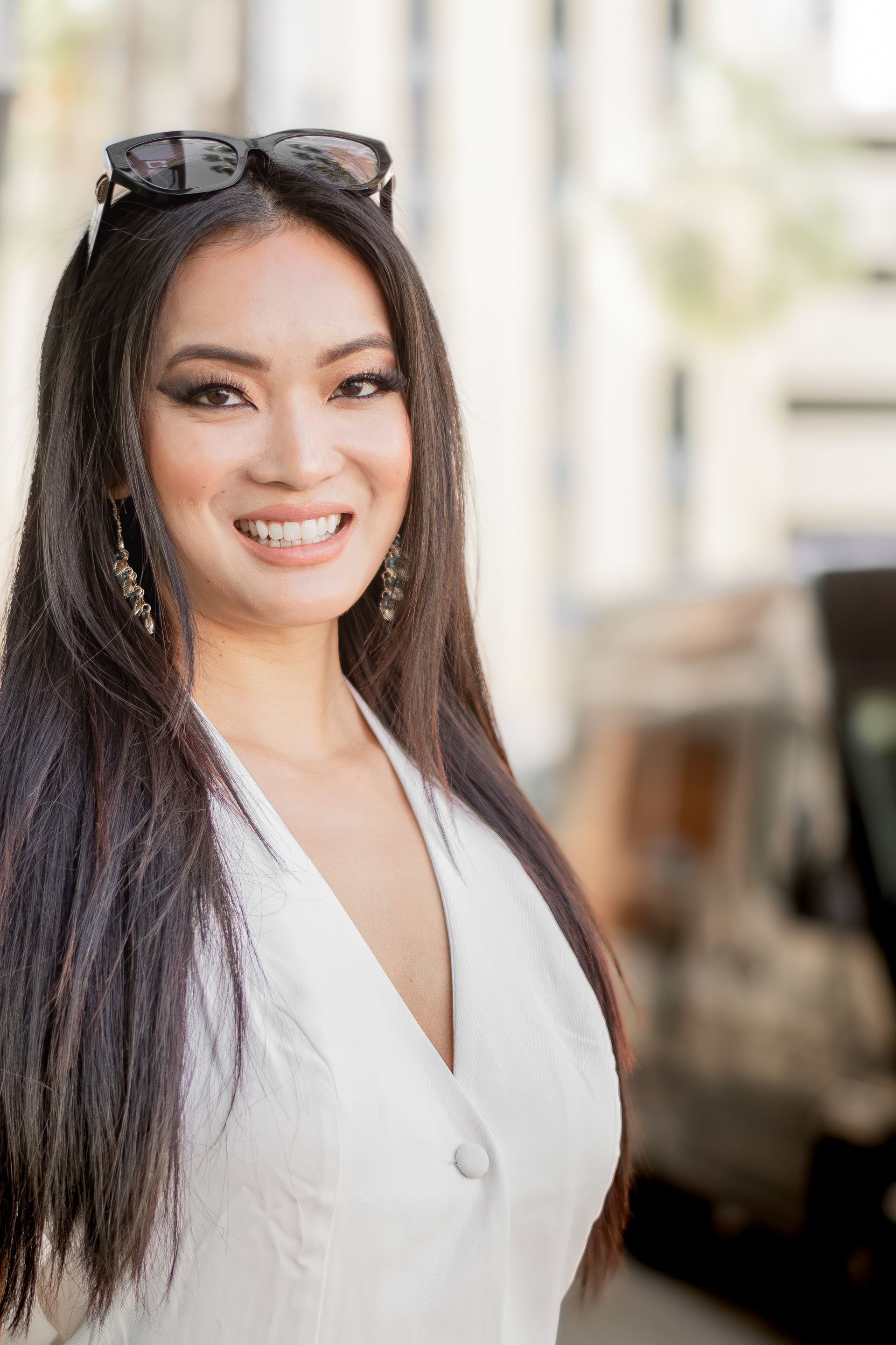 Smiling woman in a white top with sunglasses on her head, standing outdoors on a bright street.