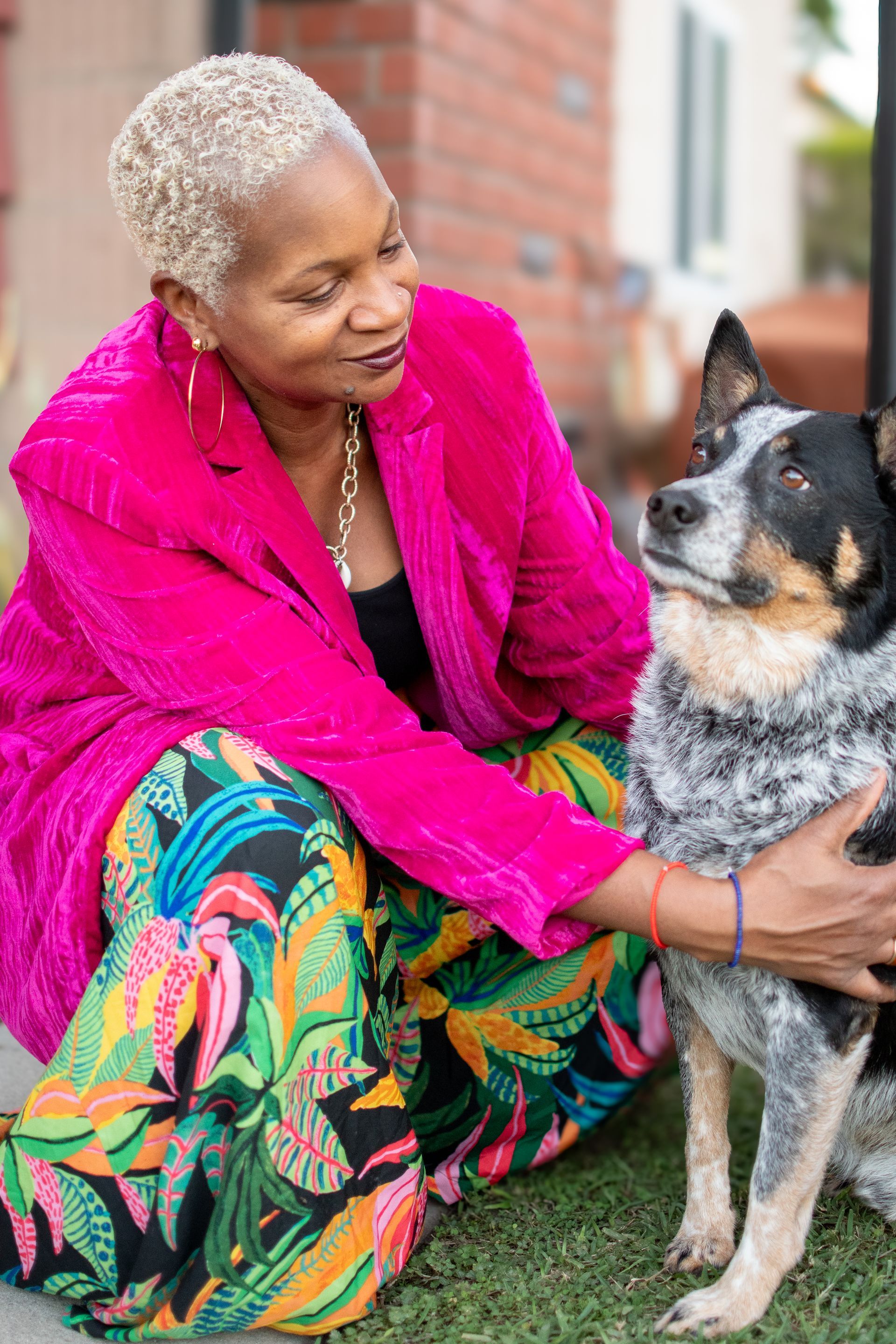 A person in a bright pink blazer and floral pants kneeling to pet a blue heeler dog outdoors.