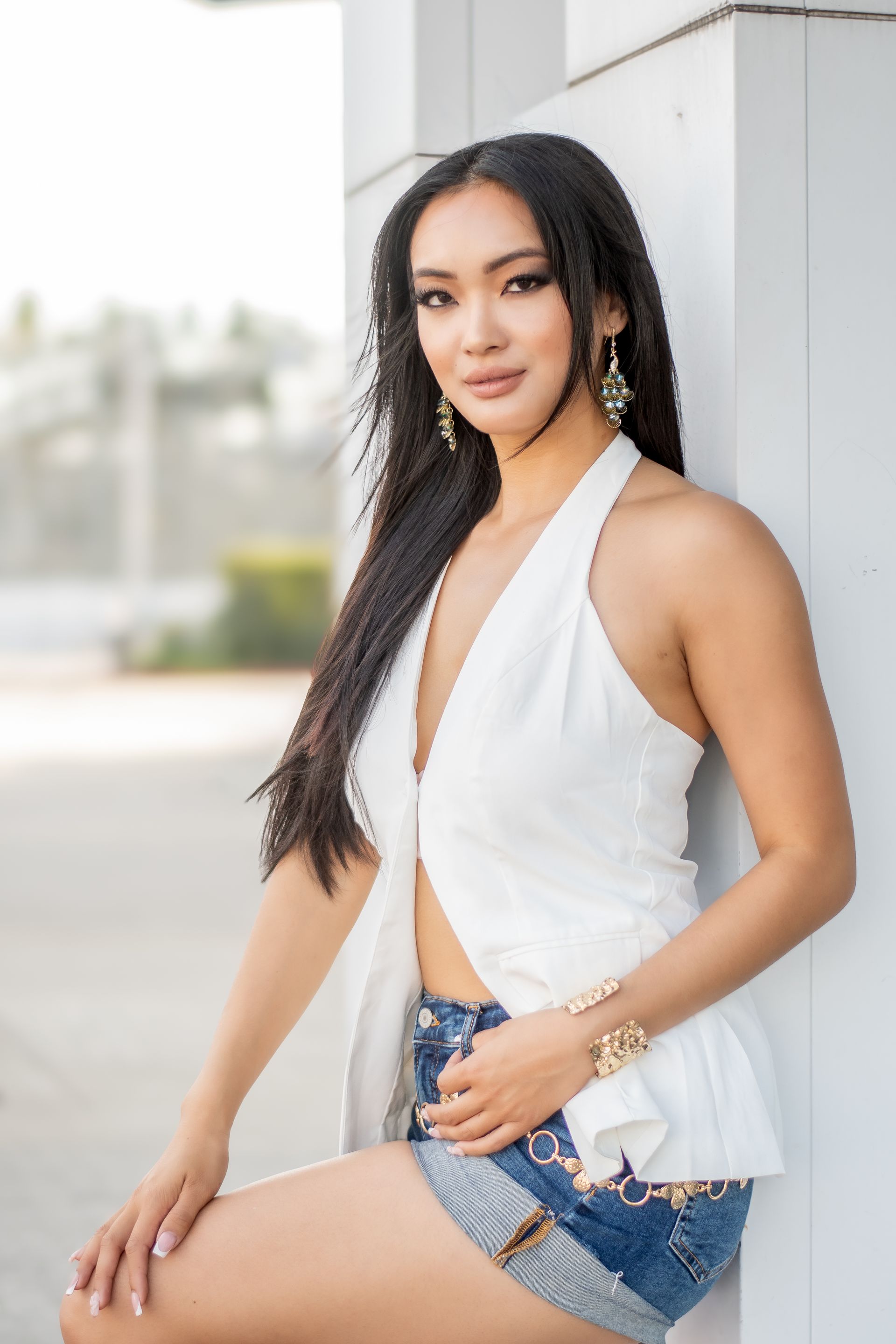 Woman in a white halter top and denim shorts leaning against a wall outdoors.