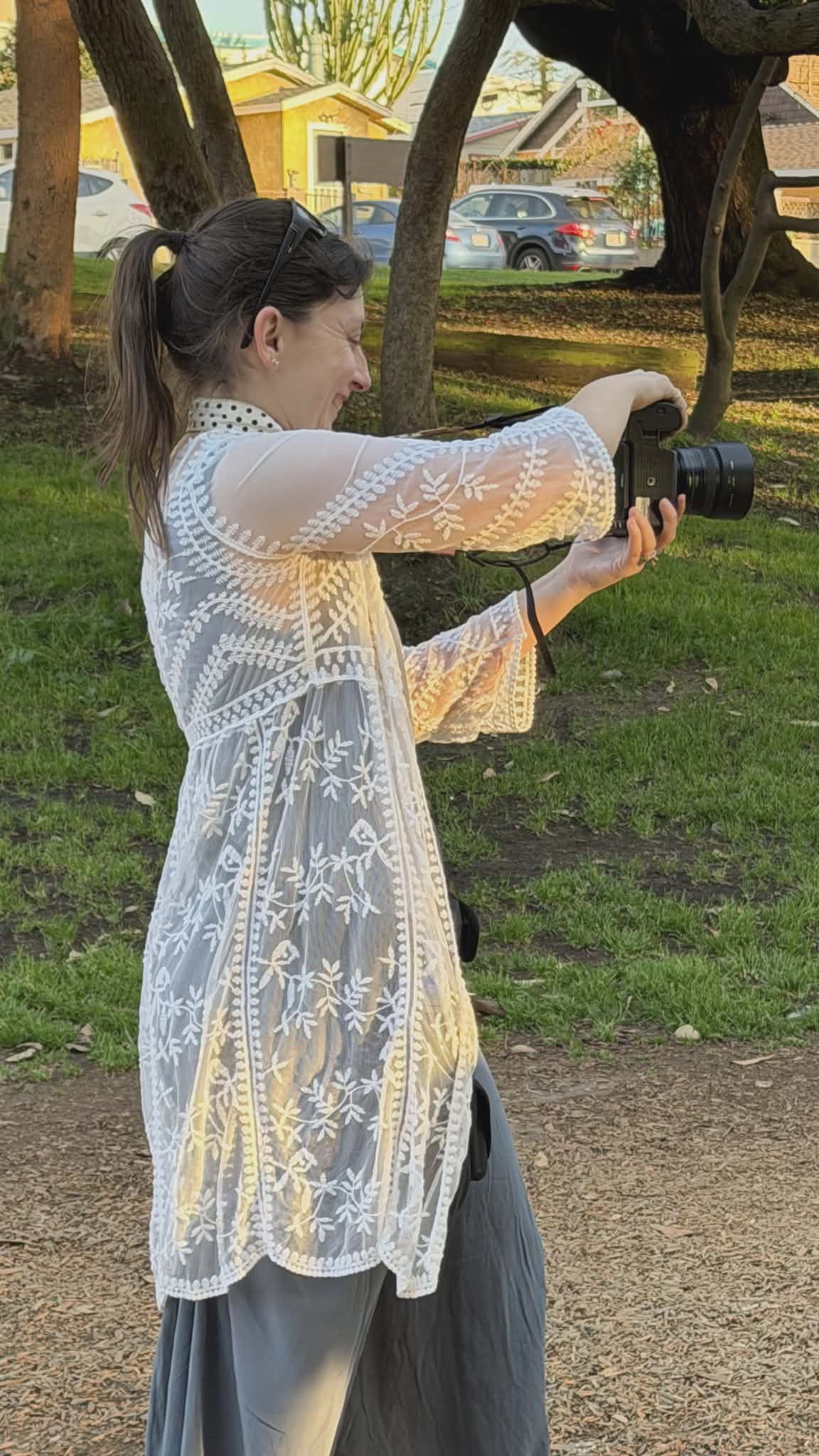 Woman taking a photo outdoors, holding a camera. She wears a white lace top and jeans, with trees and cars in the background.