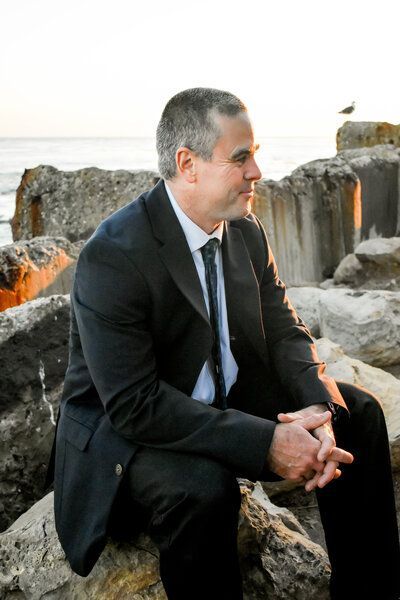 A man in a suit and tie is sitting on a rock near the ocean.