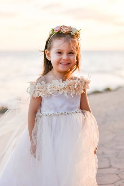 A little girl in a white dress and flower crown is standing on a beach.