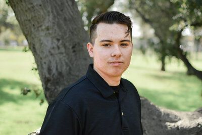 A young man in a black shirt is standing in front of a tree in a park. Senior at Banning Park