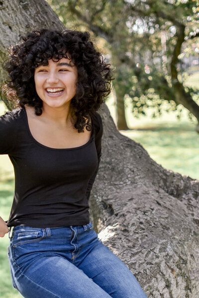 A woman with curly hair is sitting on a tree branch. Senior at Banning Park