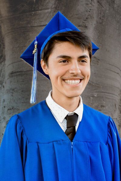 A young man wearing a blue graduation cap and gown is smiling.