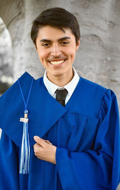 A young man in a blue graduation cap and gown is smiling and holding a tassel.