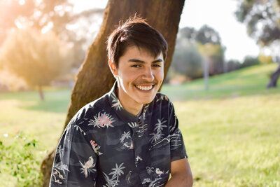 A young man is sitting under a tree in a park and smiling.
