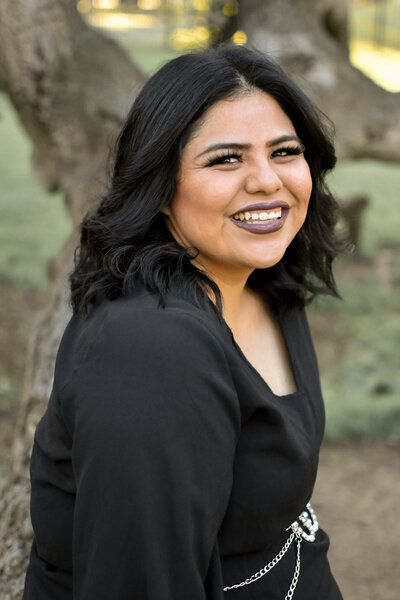A woman in a black dress is smiling in front of a tree.