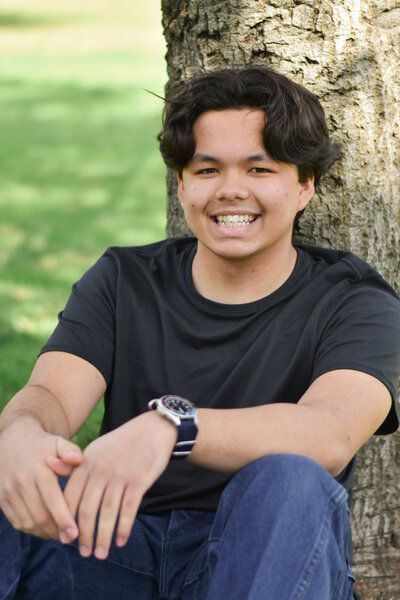 A young man is sitting under a tree wearing a watch and smiling. Senior at Banning Park