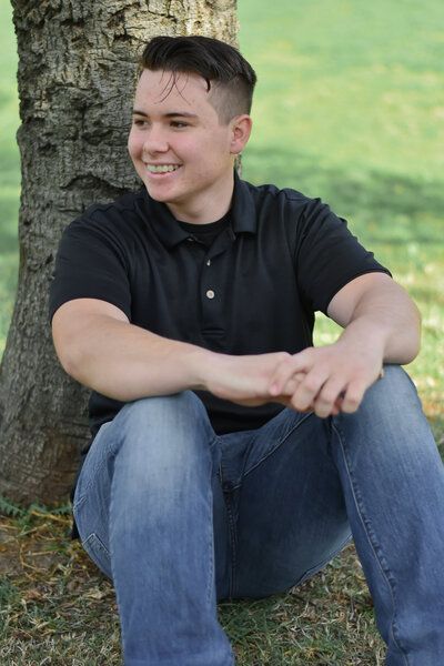 A young man is sitting under a tree and smiling. Senior at Banning Park