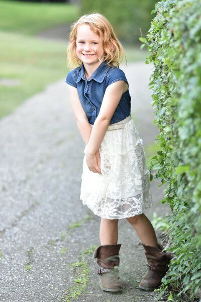 A little girl in a blue shirt and white skirt is standing next to a bush.