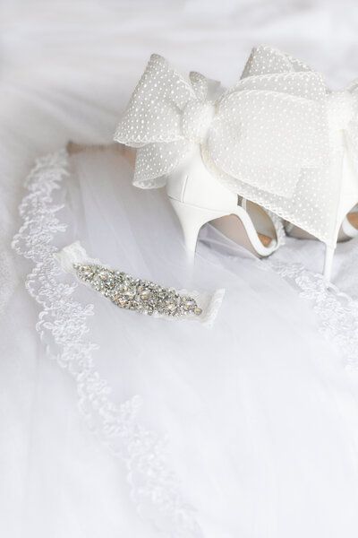 A pair of white wedding shoes with a bow on top of a white veil.