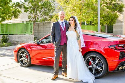 A bride and groom are posing for a picture in front of a red sports car.