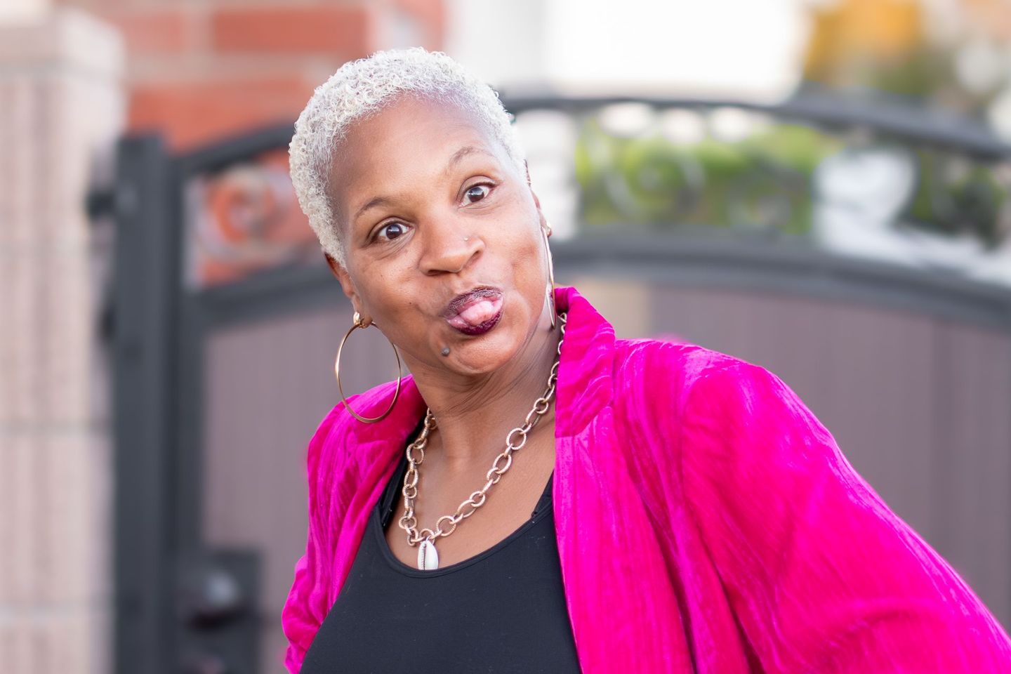 A woman with short, light-colored hair wearing a bright pink jacket makes a playful face, sticking out her tongue.