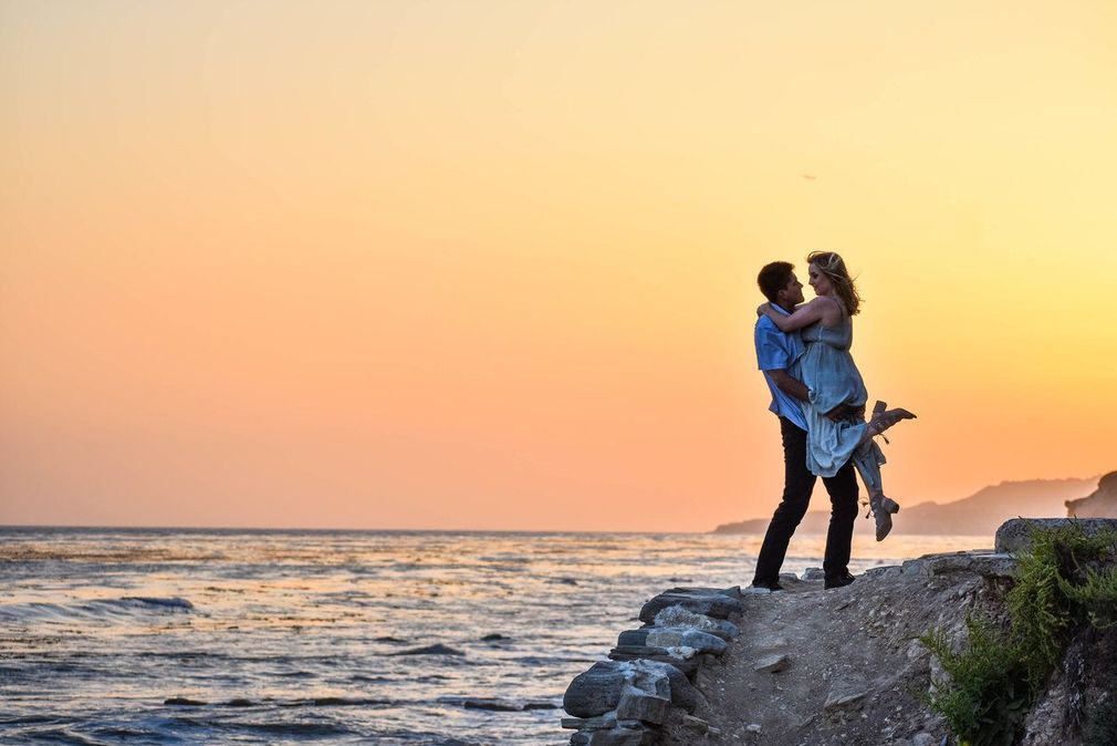 A man is holding a woman in his arms on a cliff overlooking the ocean at sunset.