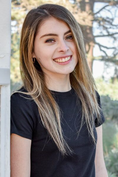 A woman in a black shirt is smiling and leaning against a wall. Senior