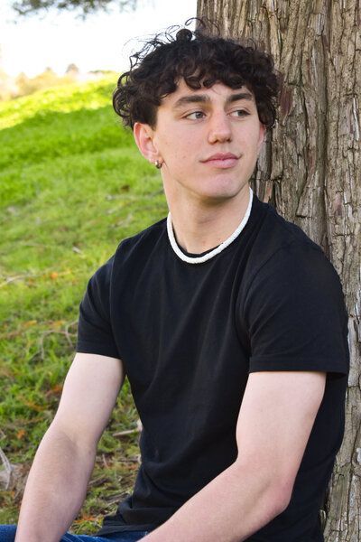A young man in a black shirt is leaning against a tree. Senior