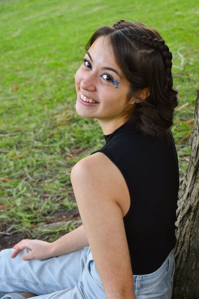 A young woman is sitting next to a tree and smiling. Senior. 