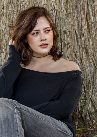 A woman in a black off the shoulder top is sitting in front of a tree. Senior