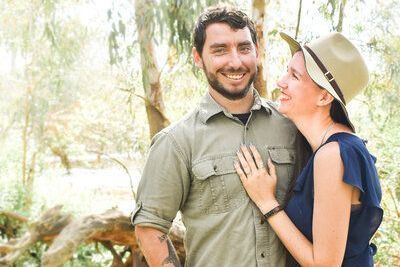 A man and a woman are posing for a picture in the woods near Huntington Library. the woman is wearing a hat.