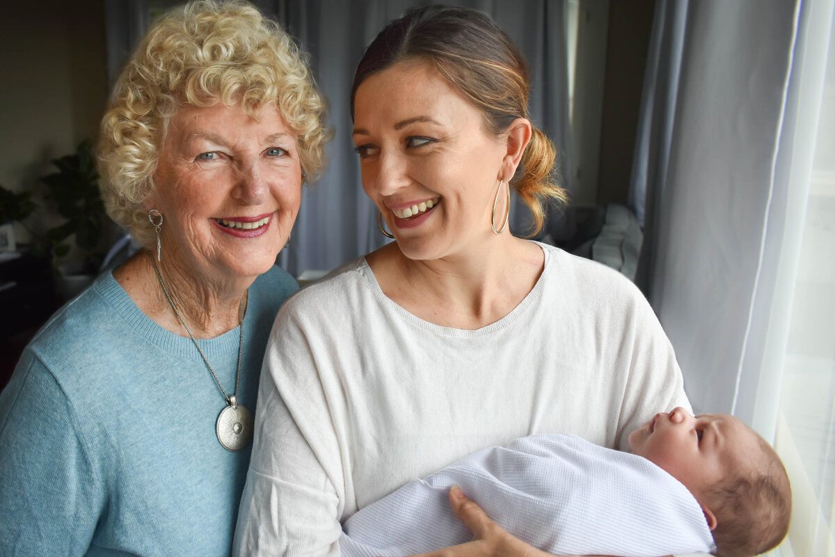 Woman holding baby, smiling with another woman. Both women near window.