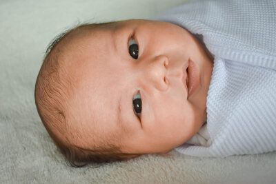 A newborn baby is wrapped in a white blanket and looking at the camera.