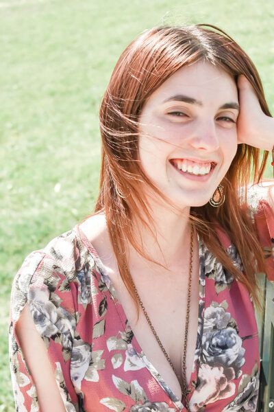 A woman in a pink floral dress is smiling while sitting in the grass.