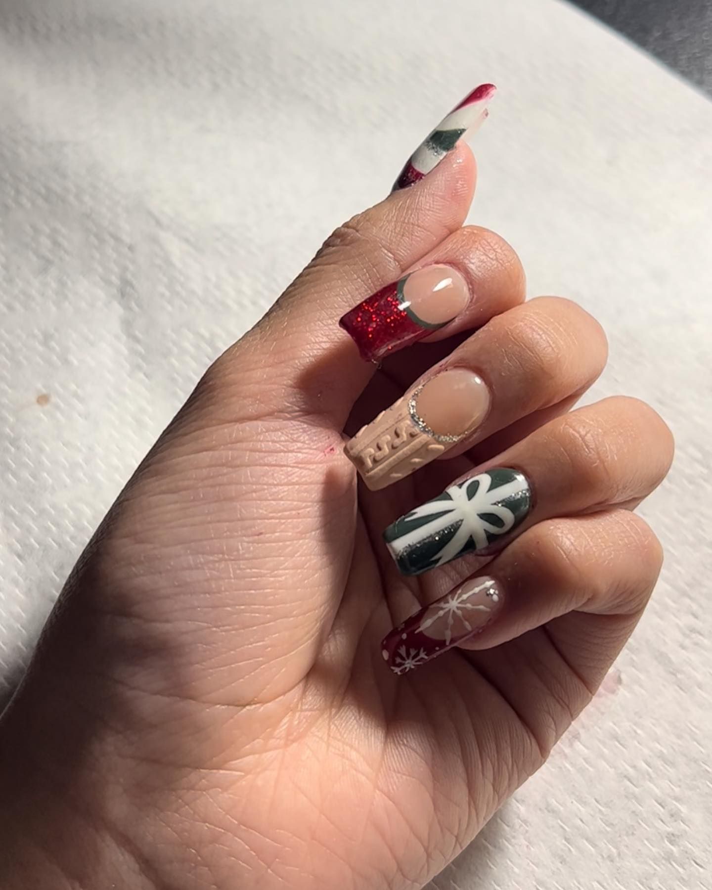 Hand with long, Christmas-themed nail art. Designs include red glitter, a gift box, and a sweater pattern.