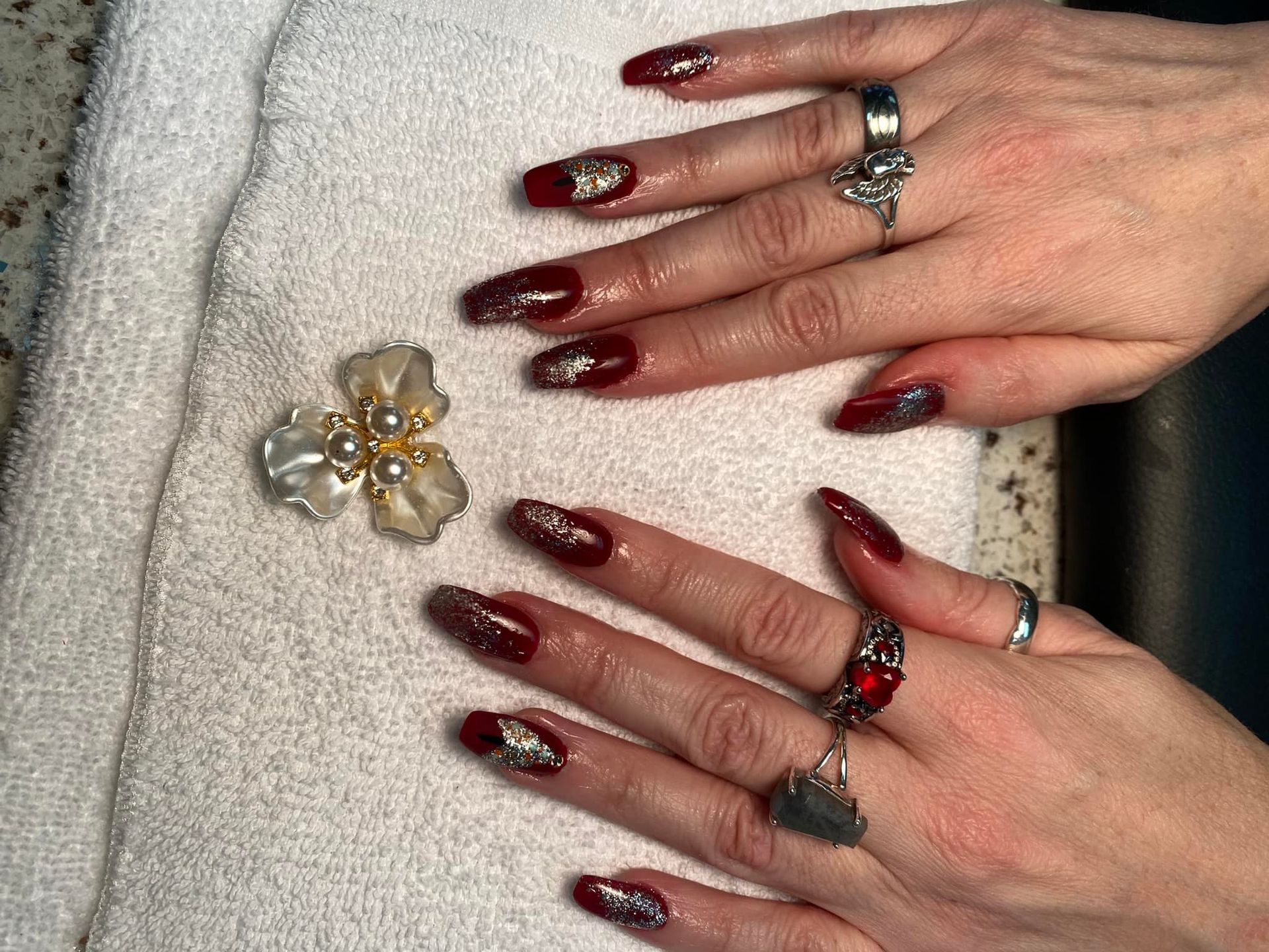 Hands with long, burgundy nails and silver glitter. Silver rings on fingers rest on a white towel, next to a flower brooch.