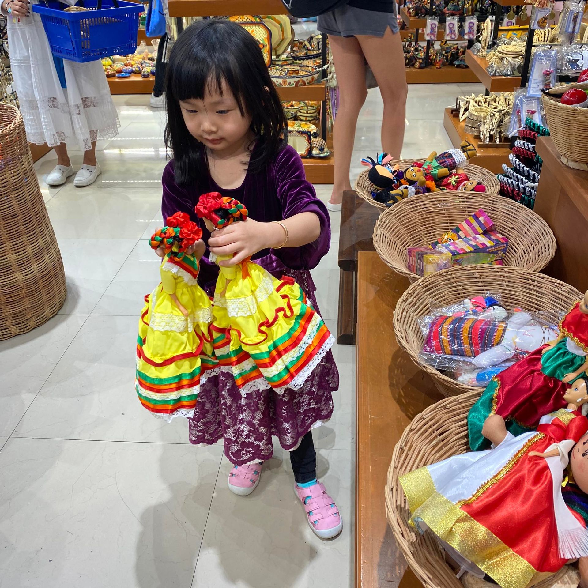 Young child holding two colorful dolls in a shop, surrounded by baskets of souvenirs.