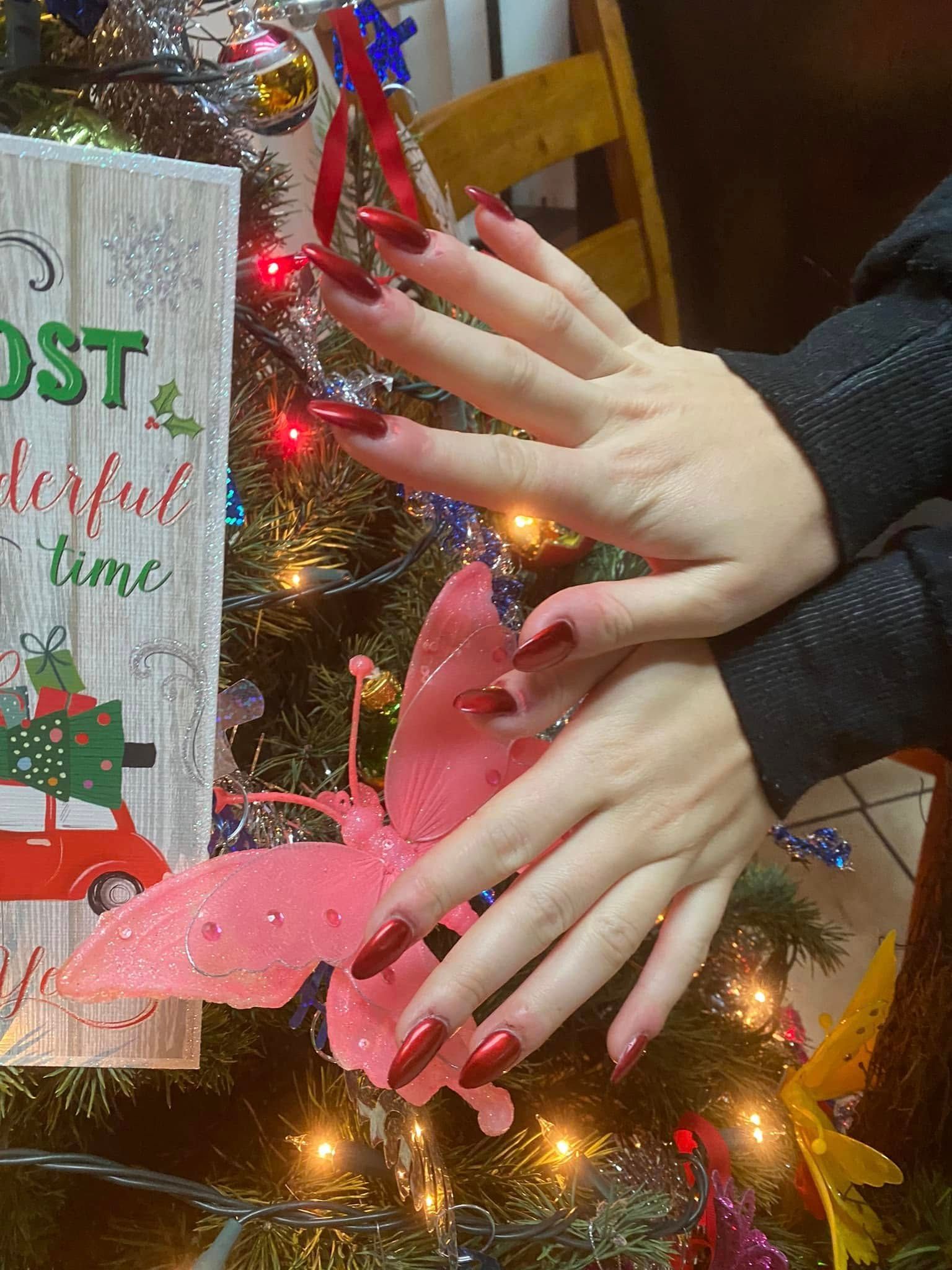 Hands with long, red nails near a Christmas tree. One hand rests on a pink butterfly ornament.