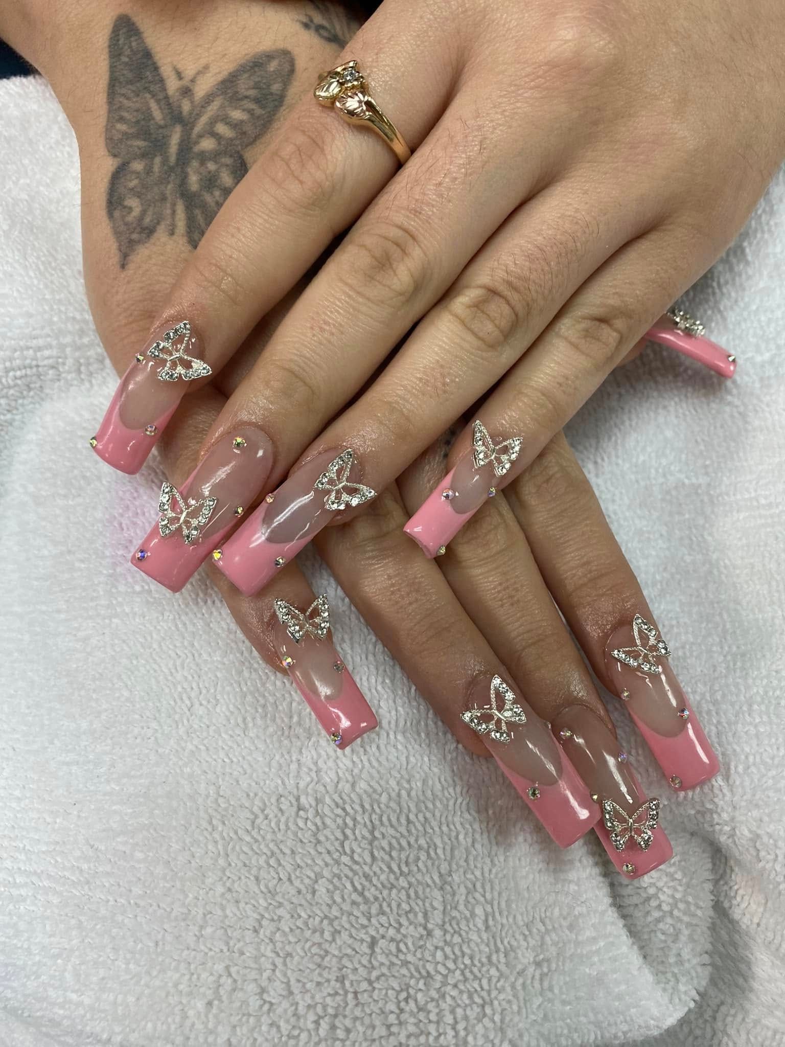 Hands with long, pink French tip acrylic nails decorated with butterfly-shaped rhinestones. Butterfly tattoo on one wrist.