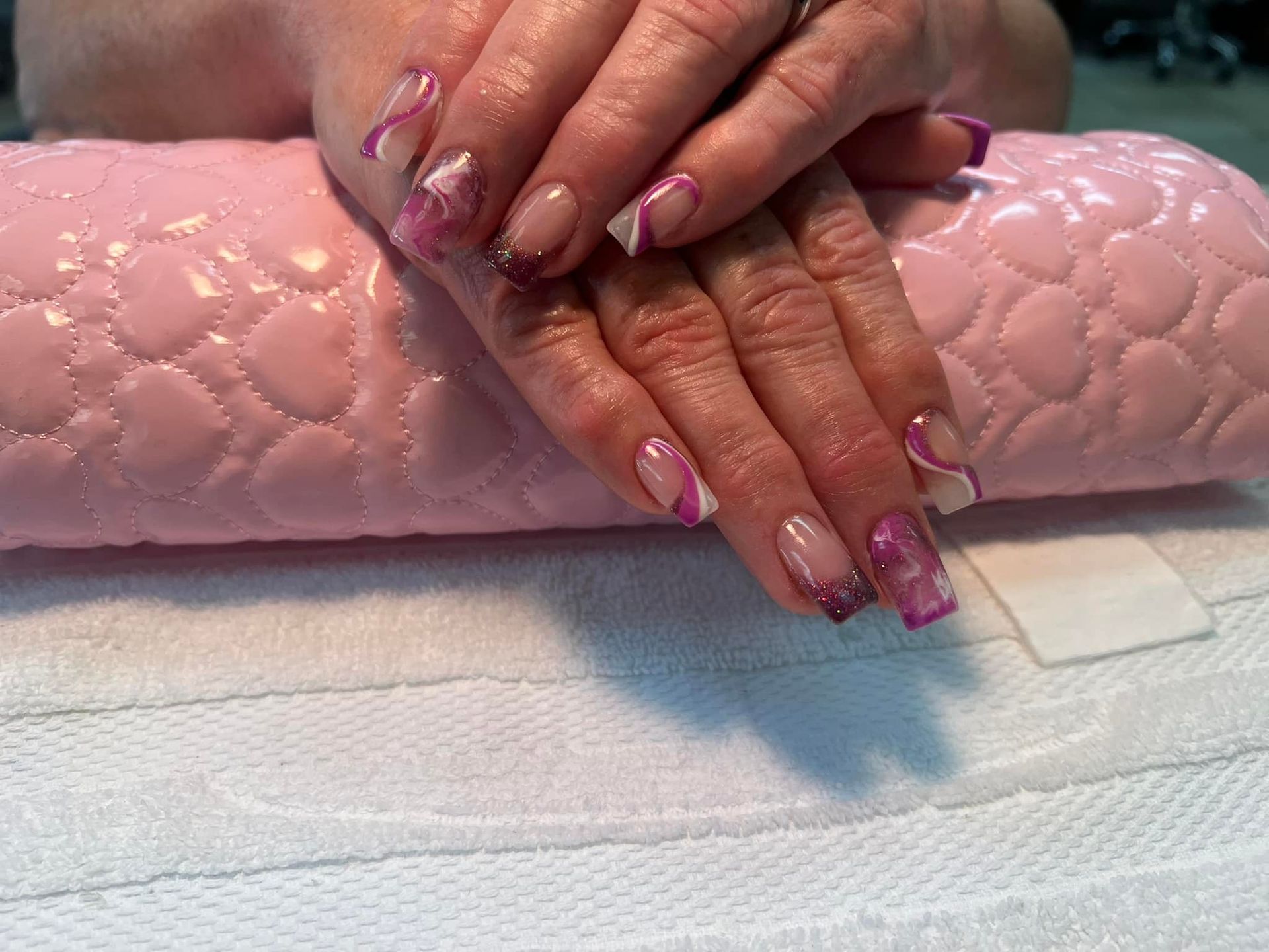 Hands with pink and white nail art, resting on a pink cushion and white towel.