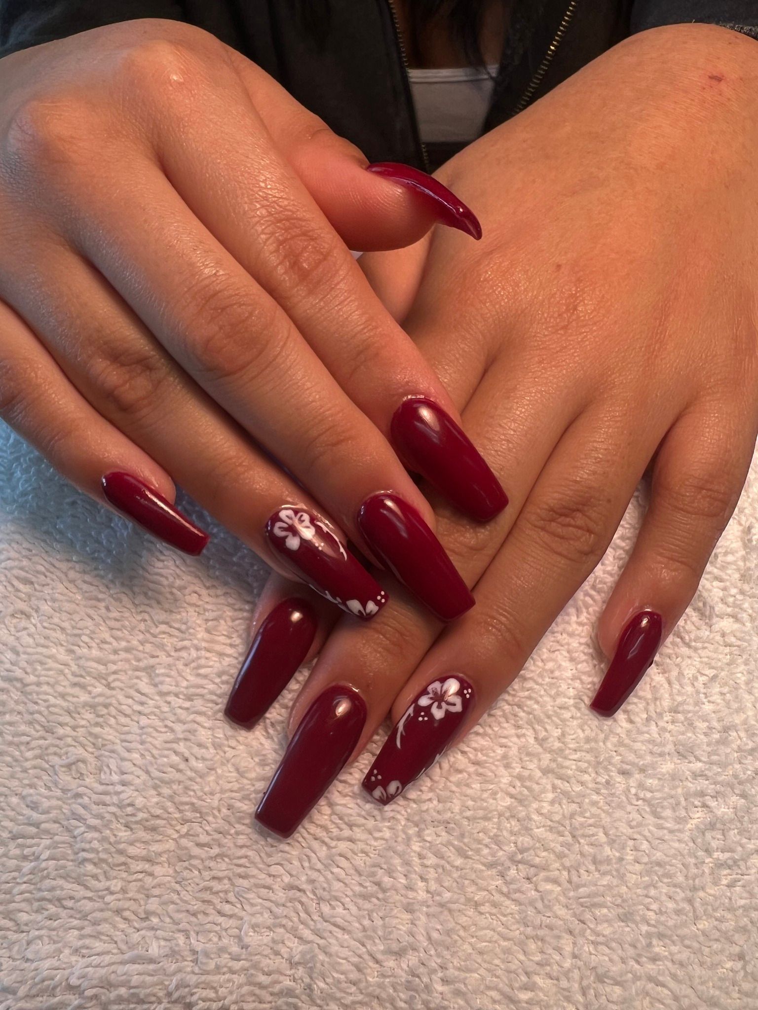 Hands with long, burgundy coffin-shaped nails, two accented with white floral designs, resting on a white towel.
