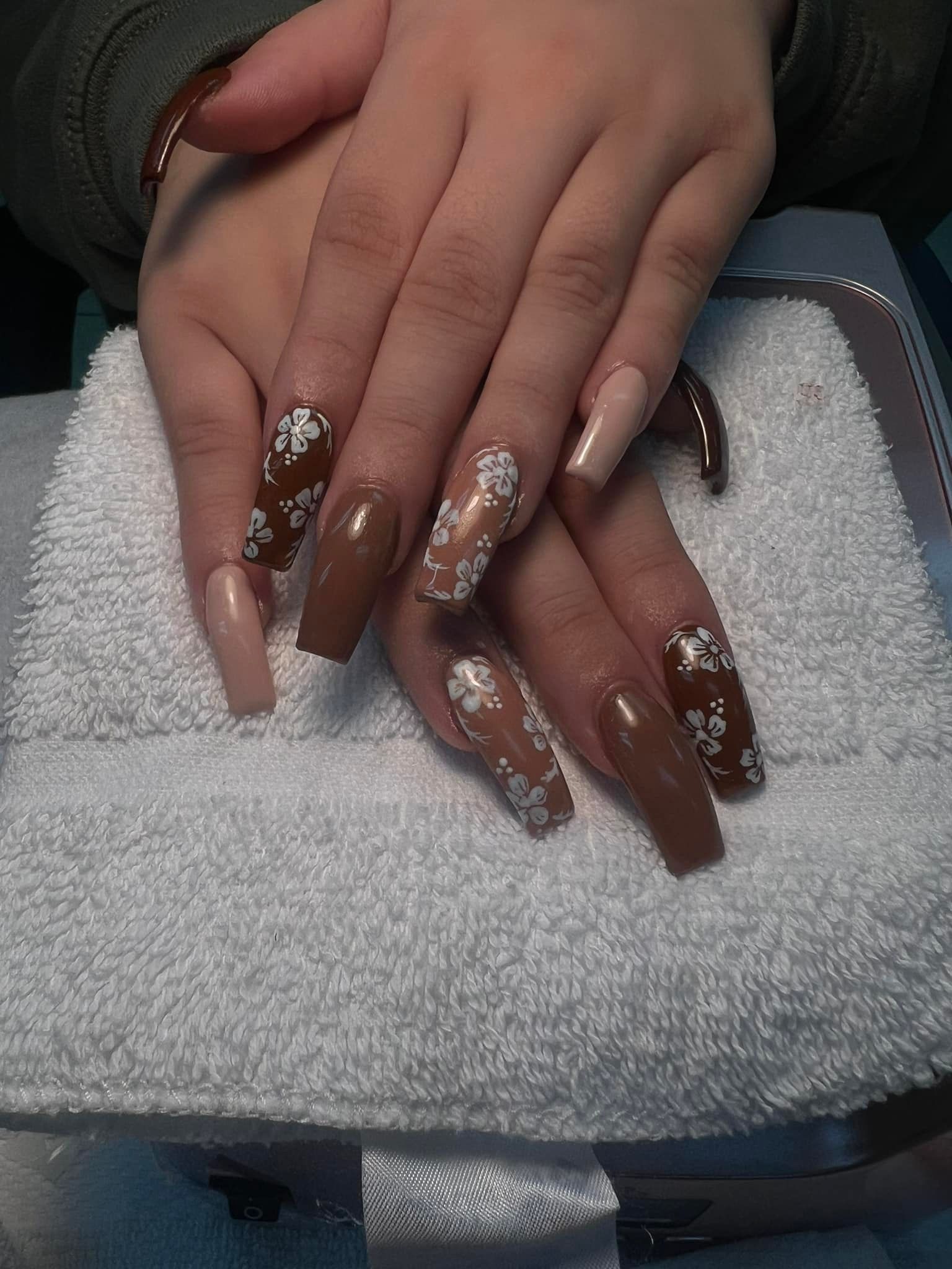 Hands with long, coffin-shaped nails. Brown and nude colors, some nails have white snowflake designs, resting on a white towel.