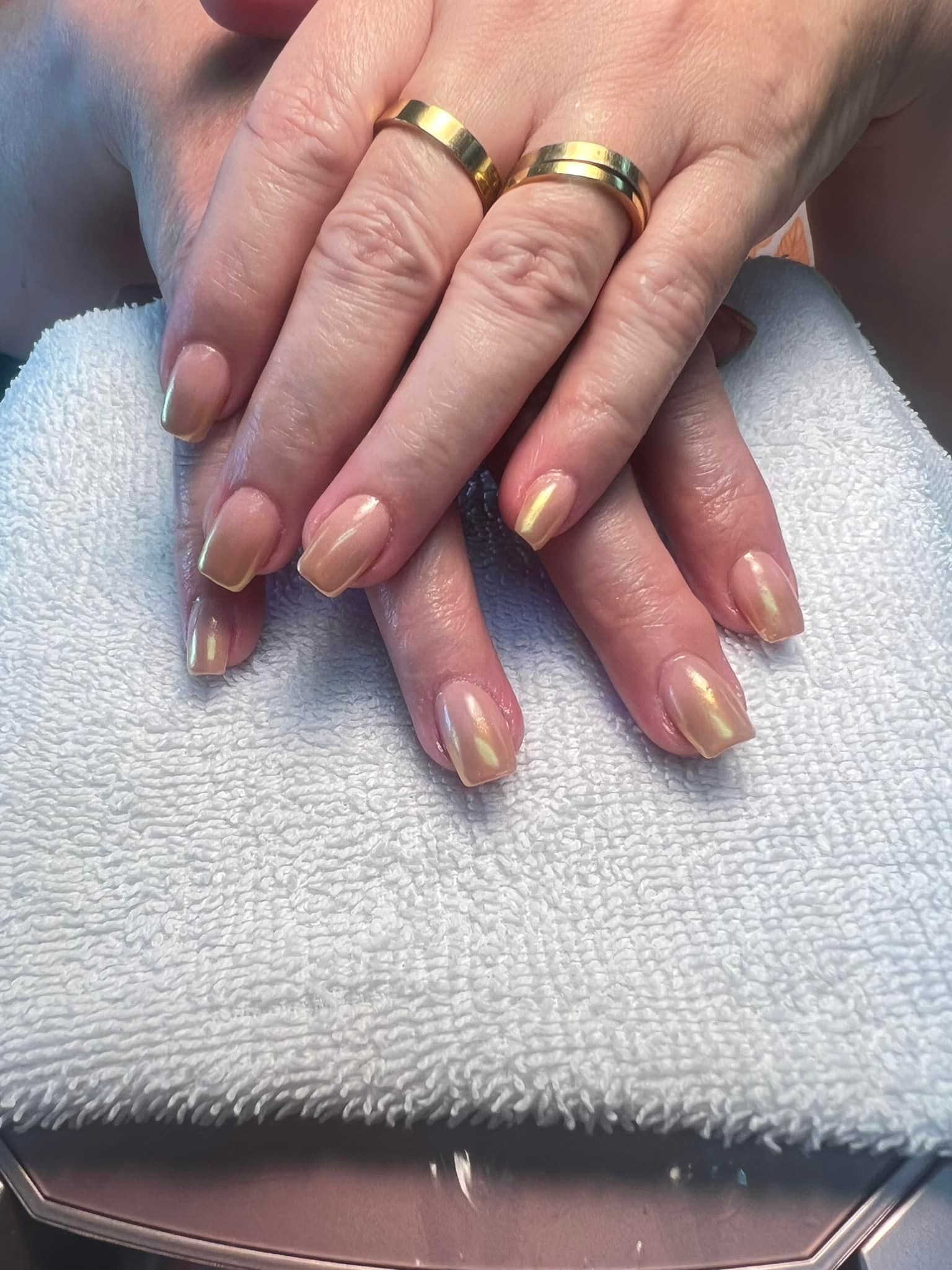 Hands with almond-shaped nails painted nude with gold accents, resting on a white towel, wearing gold rings.