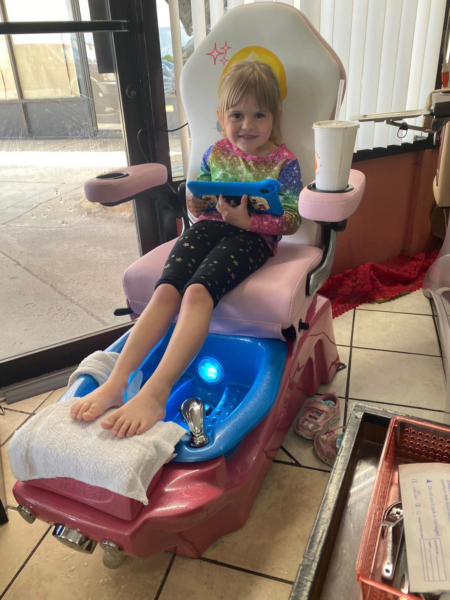 Young girl getting a pedicure at a salon, seated in a pink chair, holding a tablet.
