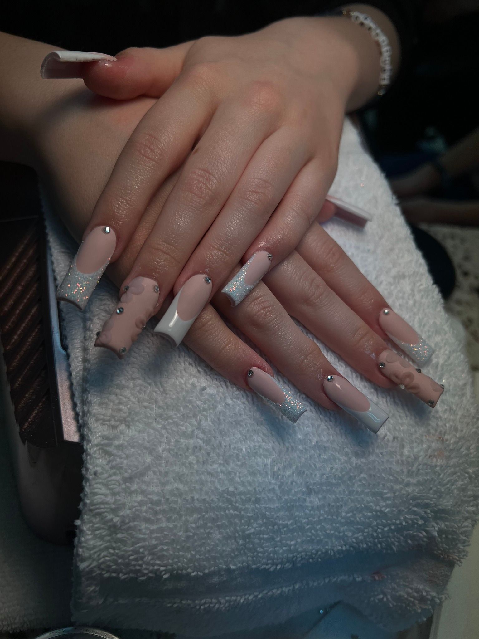 Hands with long, square-shaped acrylic nails. The nails are light pink with white tips and sparkling accents, resting on a white towel.