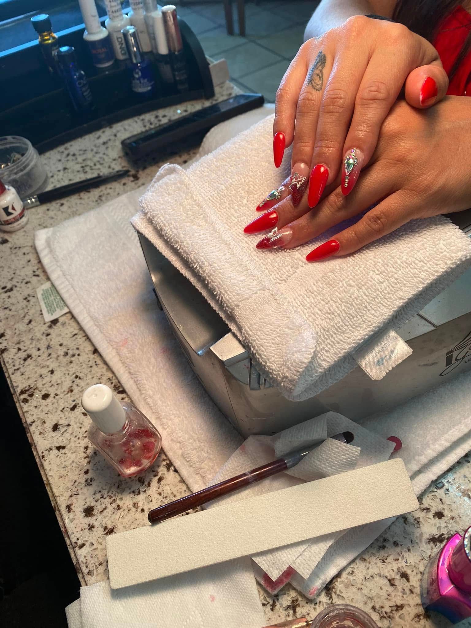 Red acrylic nails with rhinestones on a hand in a salon setting.