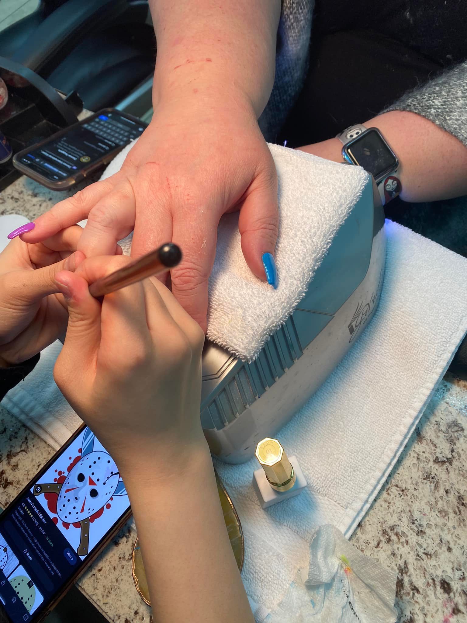 Person getting their nails painted at a salon.  A hand holds a nail brush.  Blue polish on some nails. Phone on table.