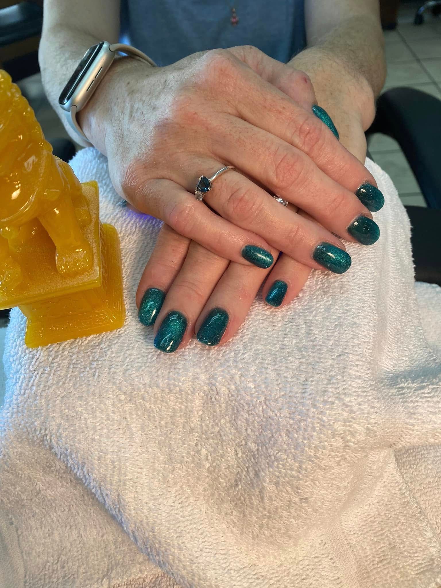 Hands with teal glitter nail polish resting on a white towel, a ring and watch visible.