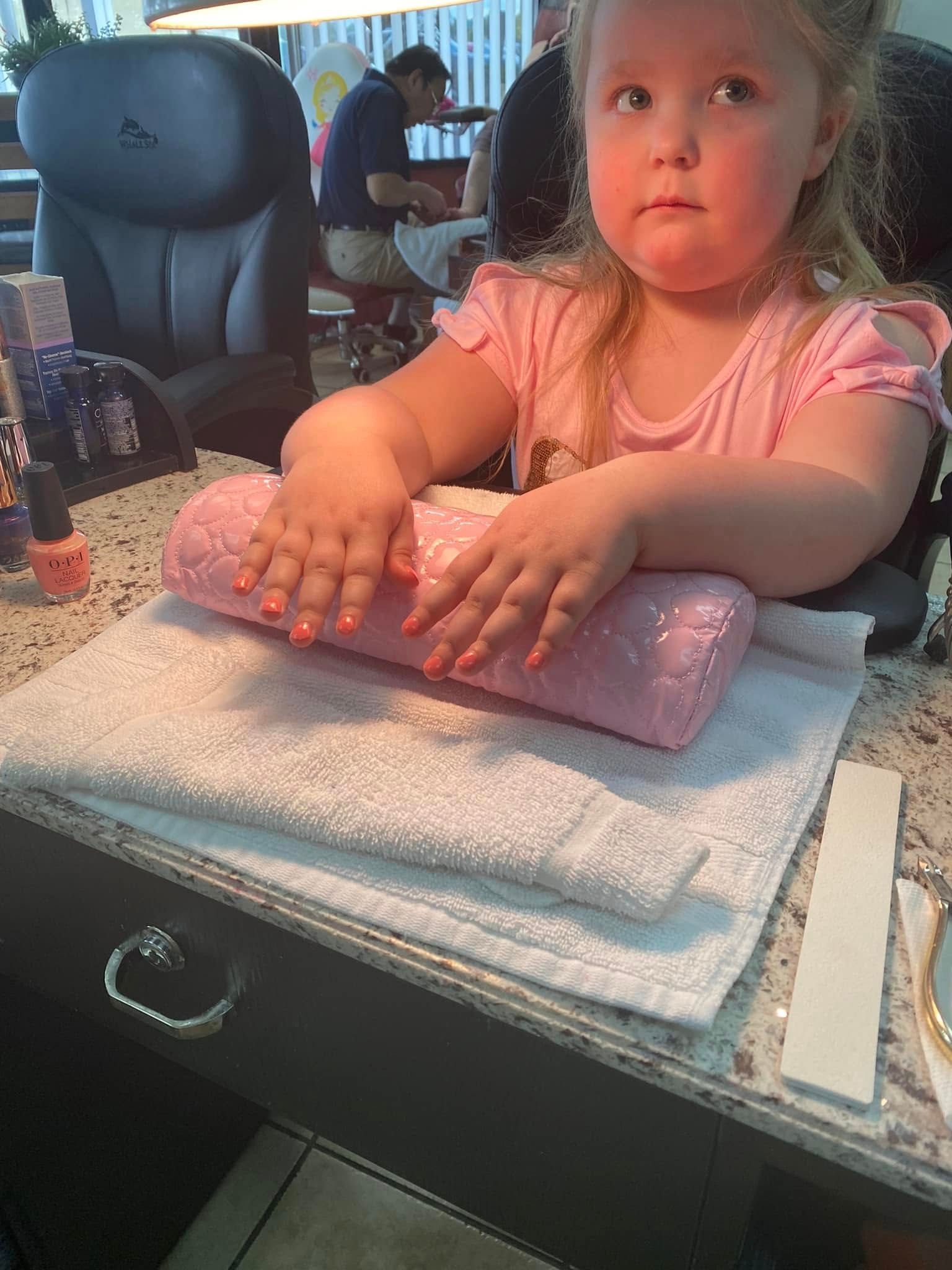 Young child getting a manicure at a nail salon, wearing pink, looking at her nails.