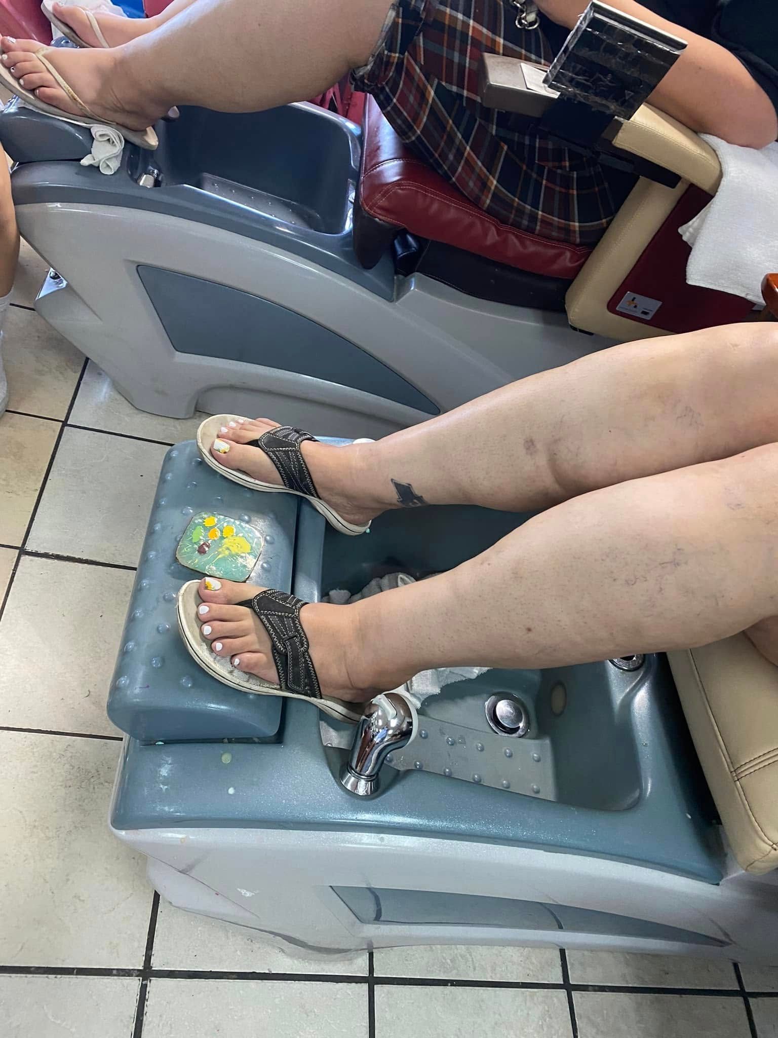 Feet in a pedicure spa, wearing flip-flops. One leg has a tattoo, light skin, and painted toenails.